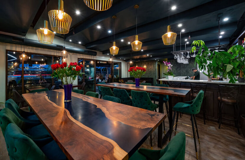 Bayshore Lounge interior dining area with wood tables, green chairs, floral centerpieces, and warm pendant lighting.